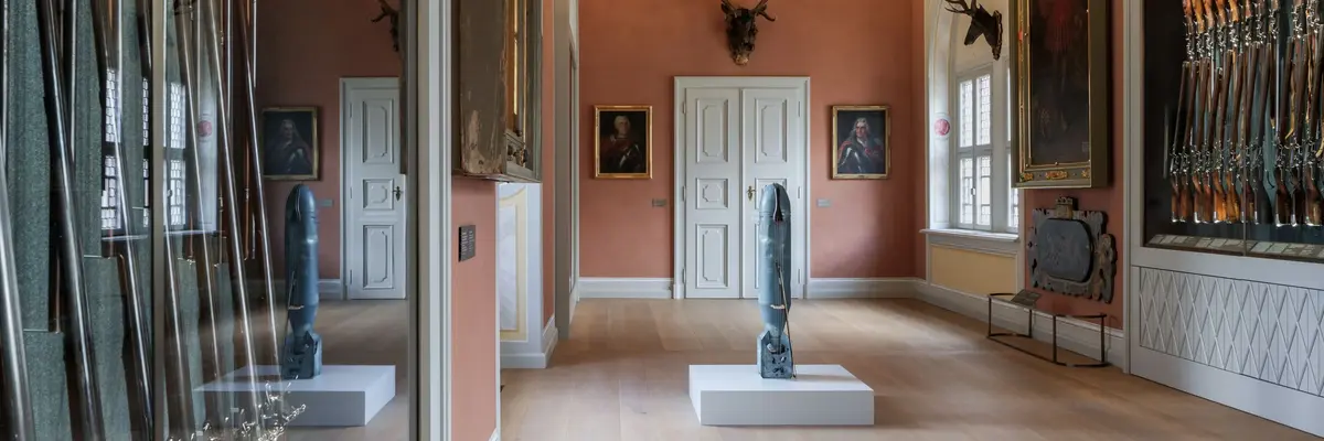 Eine zum Cello umgebaute Bombe der Künstlerin Charlotte Moorman, steht im Langen Gang umgeben von mit vielen Schusswaffen gefüllten Vitrinen.